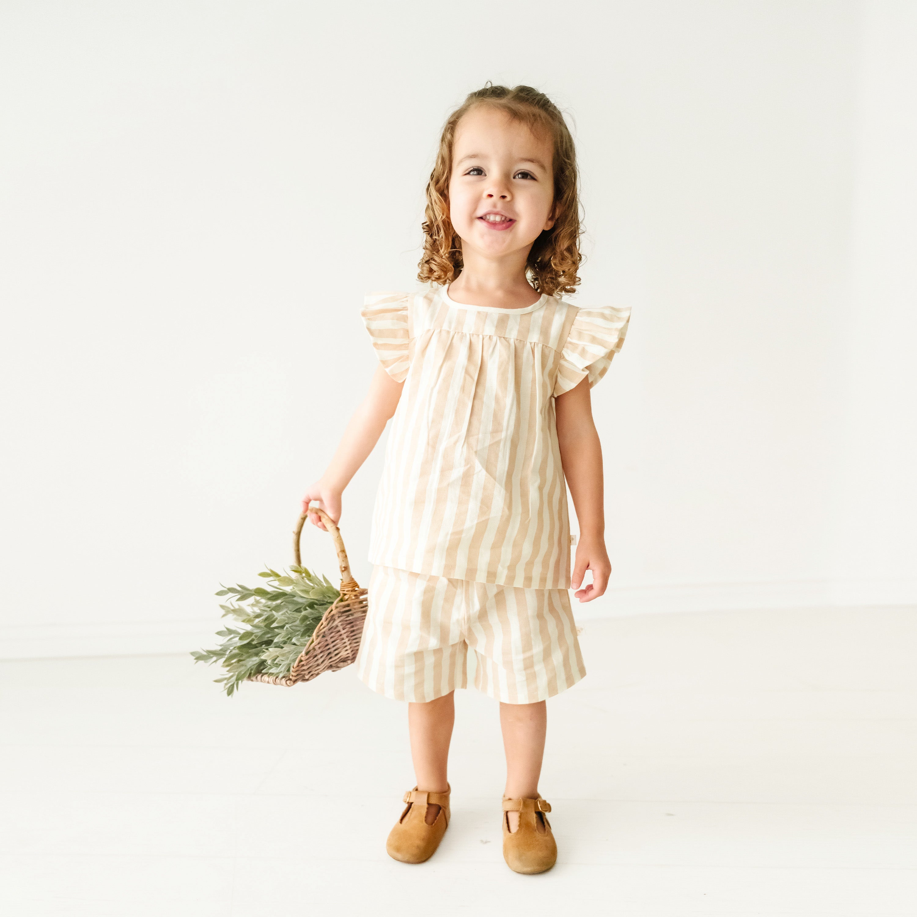 A young girl with curly hair, dressed in a Makemake Organics Organic Linen Flutter Top and Shorts - Beige Stripes, stands smiling and holding a wicker basket with greenery, on a plain white background.