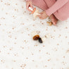 A child in pink pants sits on a floral blanket, playing with Makemake Organics' wooden toys including mushrooms and a small rolling pin.