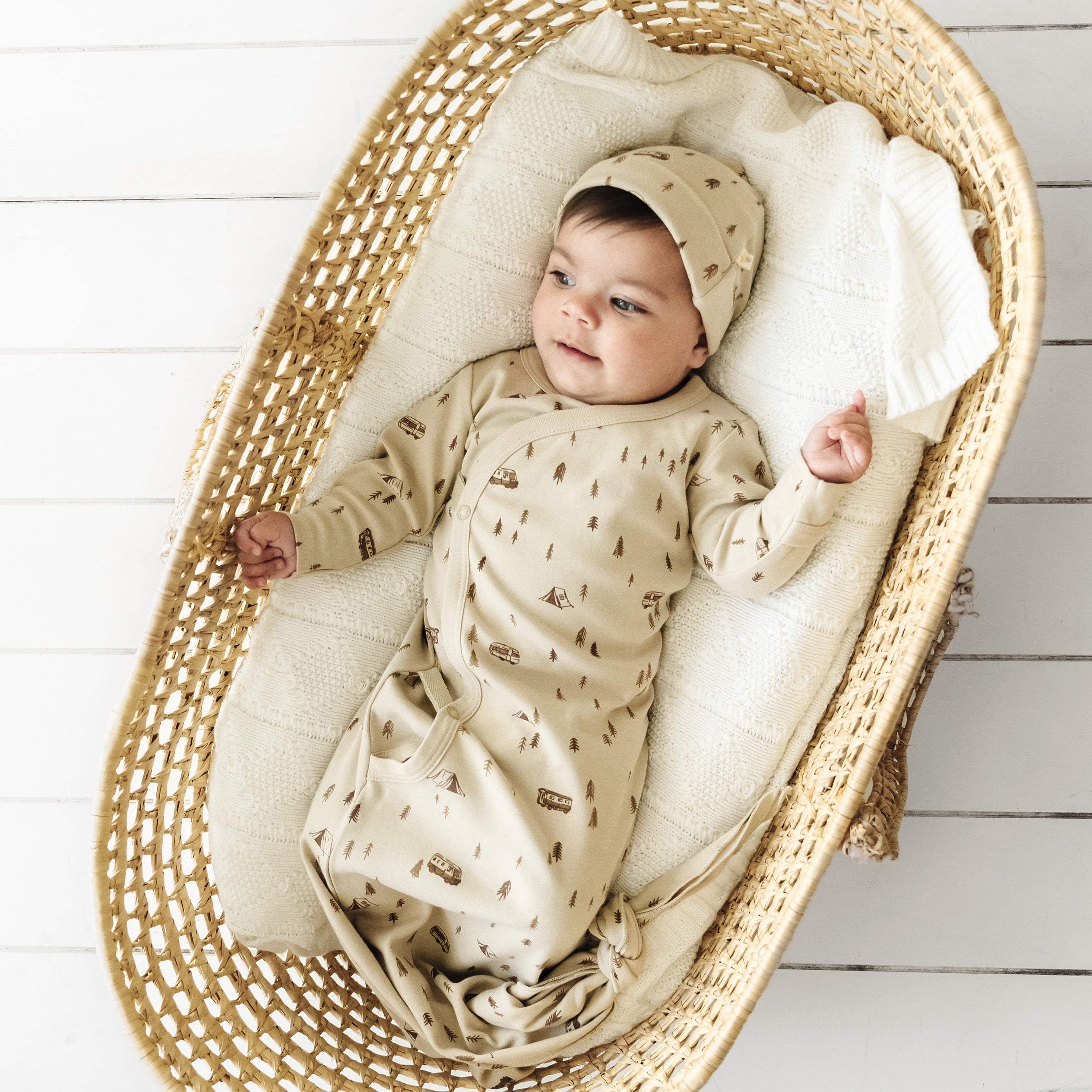 A baby in a beige Organic Baby Kimono Knotted Sleep Gown - Camplife with a hat lies comfortably inside a woven basket, placed on a white wooden floor. The onesie is adorned with tiny teepee patterns.