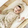A baby with wide eyes lies in a wicker basket, wearing an Organic Baby Kimono Knotted Sleep Gown in Camplife adorned with tiny woodland prints and a matching hat.
