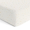 A close-up of a white fabric surface with a pattern of small, multicolored speckles including yellow, red, blue, and green, focusing on the neat corner seam of the Makemake Organics Organic Cotton Sheet Set - Dotty.