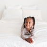 A young girl with a joyful expression lying prone on her elbows on a bed with Makemake Organics Organic Cotton Sheet Set - Dotty, gazing to her left.