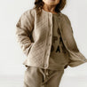 A young child wearing an Organic Kids Organic Merino Wool Buttoned Jacket in Mocha over a beige sweatshirt with "play day" printed on it, paired with matching pants, standing against a white background.