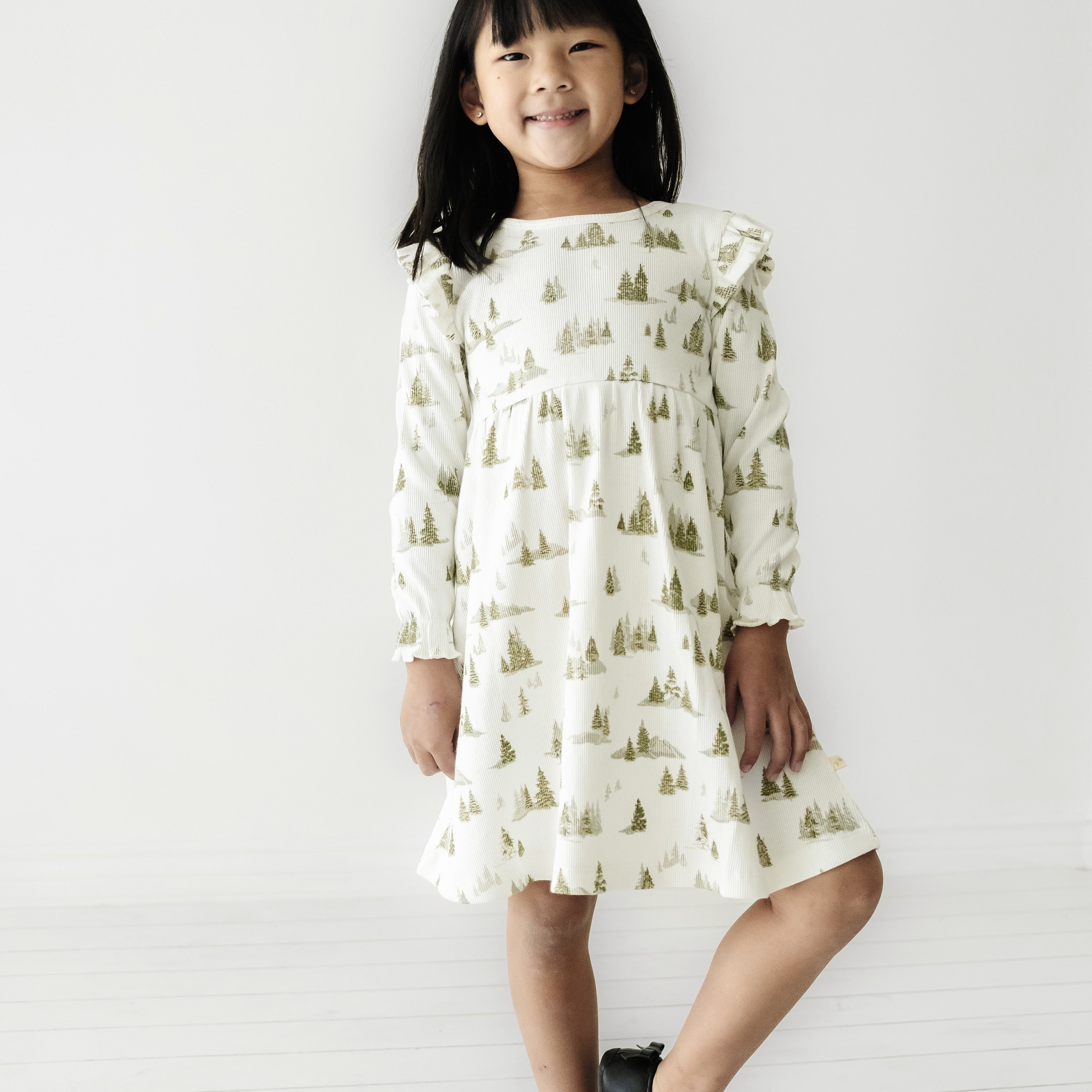 A young Asian girl smiling at the camera, wearing an Organic Girls Organic Ruffle Dress - Frosted Fir with a tree pattern, standing against a plain white background.