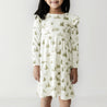A young girl stands against a plain white background, wearing a long-sleeve dress from Organic Girls adorned with a green tree pattern. She has dark hair and is smiling slightly.