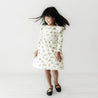 A young girl with dark hair in a playful pose, wearing an Organic Girls frosted fir dress with a tree pattern, and black shoes, spinning in a brightly lit white studio space.