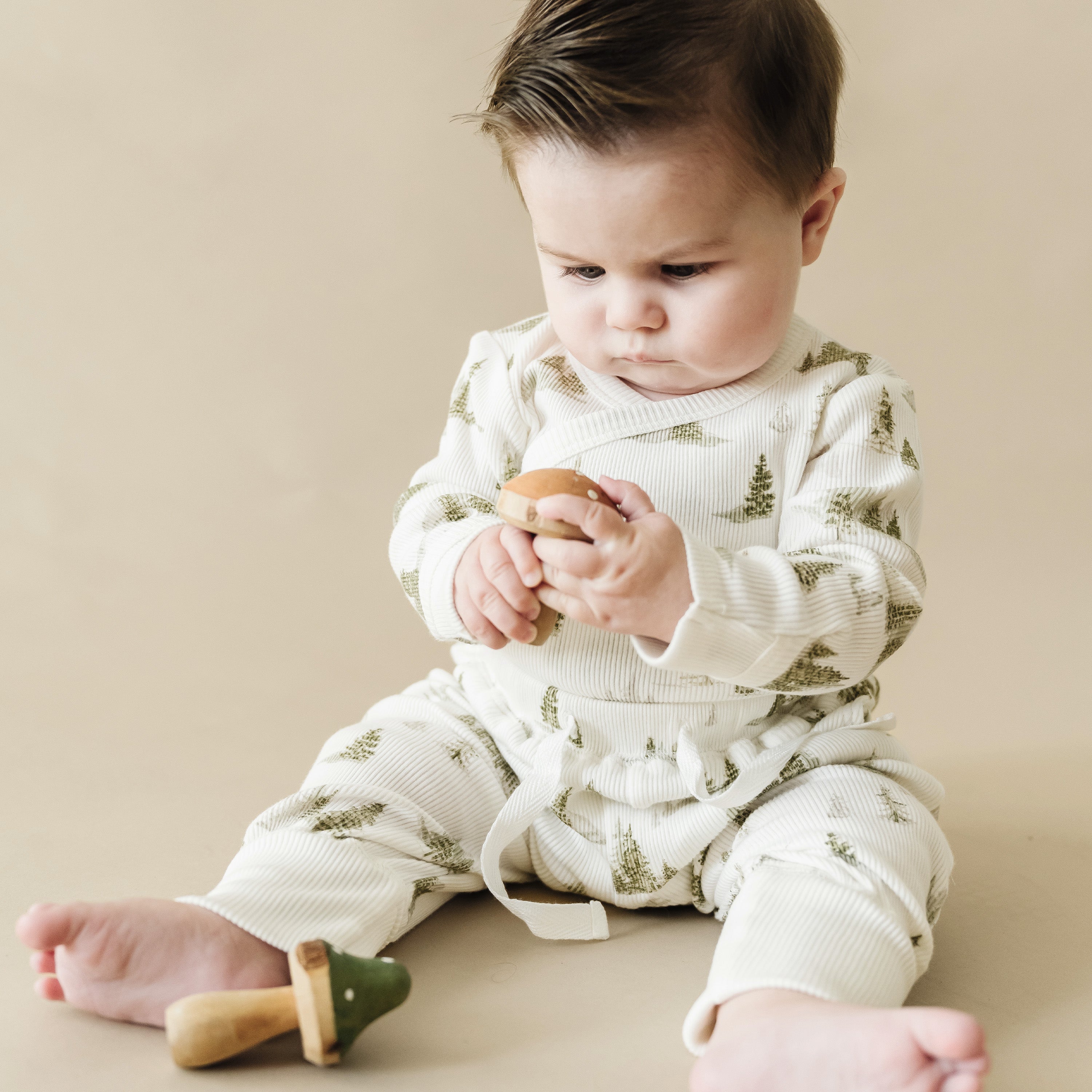A baby in a white and green Organic Baby outfit sits on a beige surface, intently examining a wooden toy, with another toy beside them.