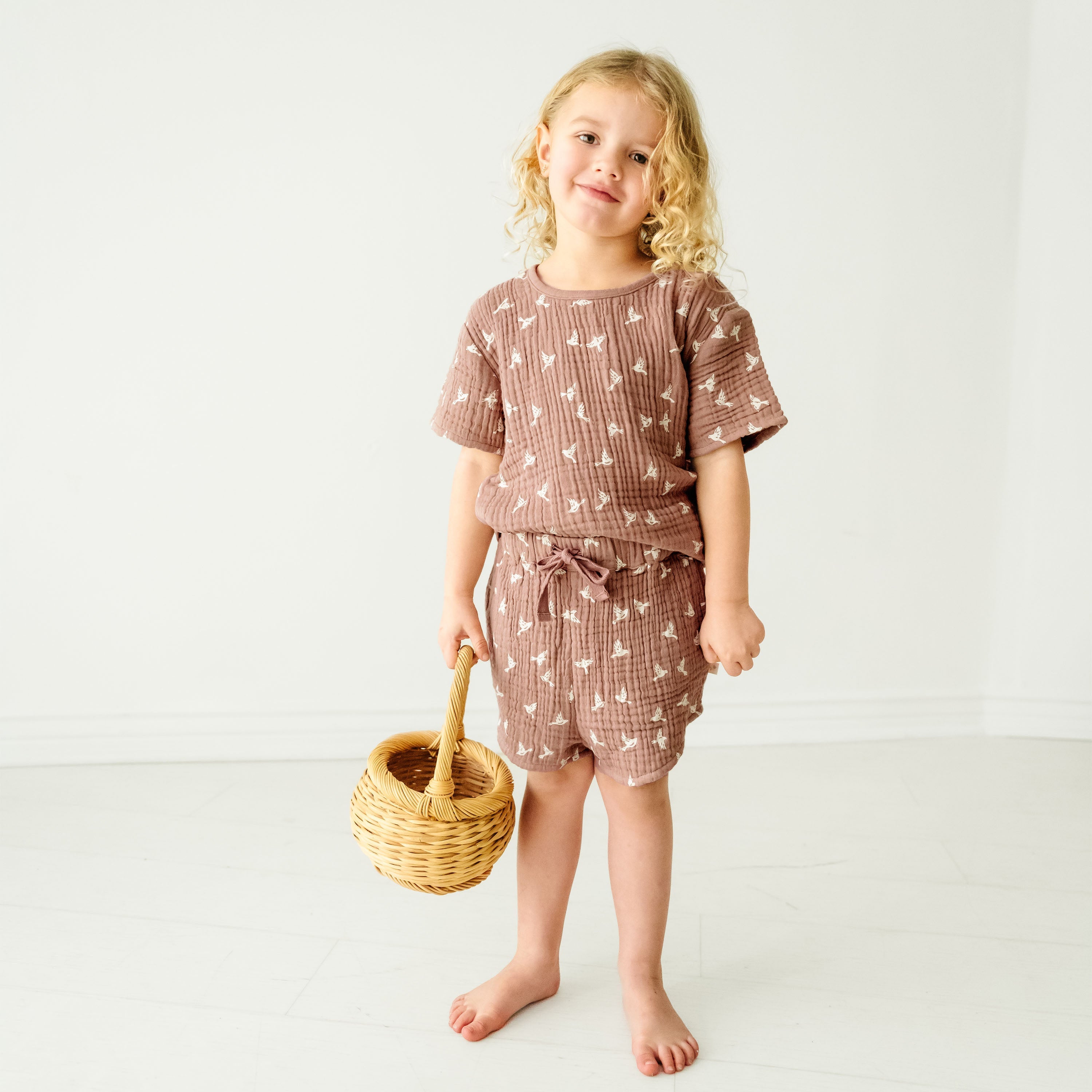 A young toddler with curly blonde hair, wearing a brown patterned outfit, stands smiling in a white room while holding a small wicker basket of Makemake Organics' Organic Muslin Top and Shorts 2 Piece Set - Flock.