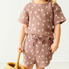 A baby in a pink, bird-patterned Makemake Organics Organic Muslin Top and Shorts 2 Piece Set - Flock holds a wicker basket, with a focus on the textured fabric and playful design of the clothes. The background is a soft, light-colored wall.