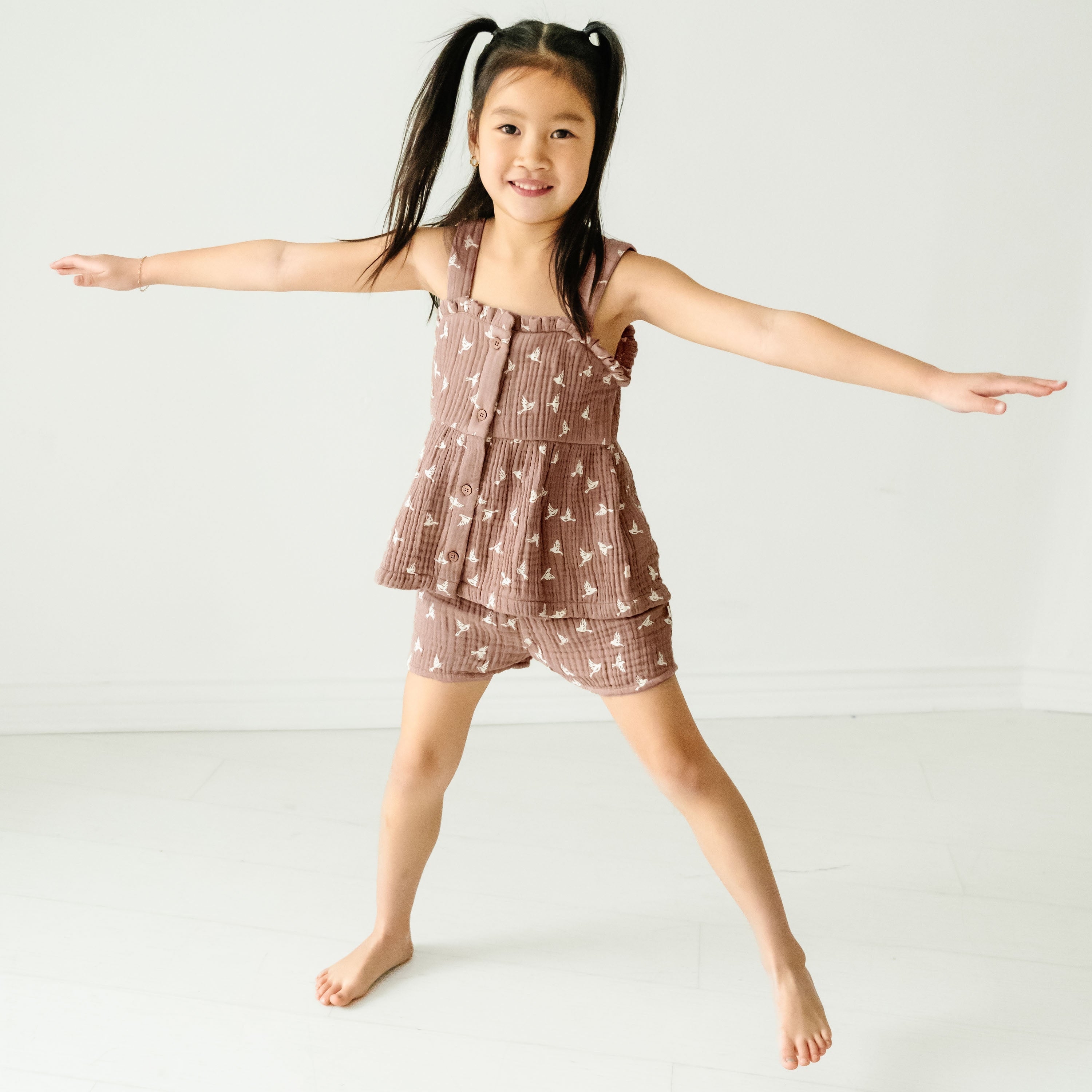 A young girl with ponytails, wearing a Makemake Organics Organic Muslin Peplum Top and Shorts Set - Flock, joyfully spreads her arms wide while standing barefoot in a brightly lit, white studio room.