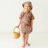A cheerful young toddler wearing a striped brown outfit and an Organic Muslin Bucket Sun Hat - Flock by Makemake Organics, holding a wicker basket, stands smiling in a bright, neutral-colored room.