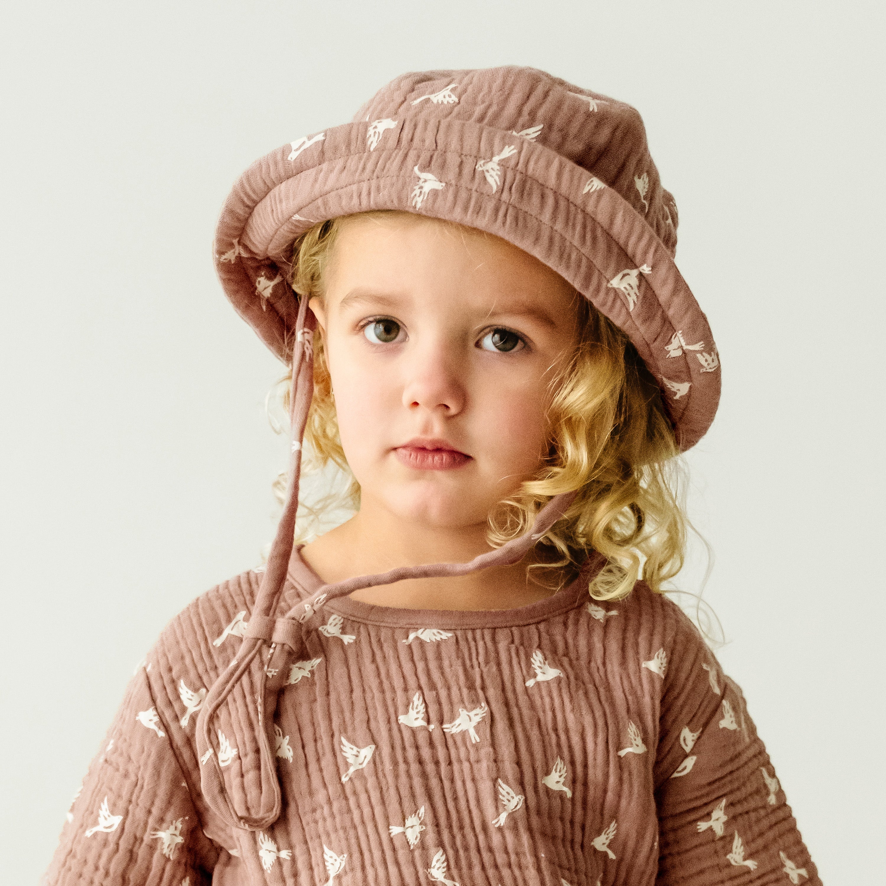 A young boy with curly blonde hair wearing a Makemake Organics Organic Muslin Bucket Sun Hat - Flock and a shirt patterned with white deer, looking thoughtfully to the side.