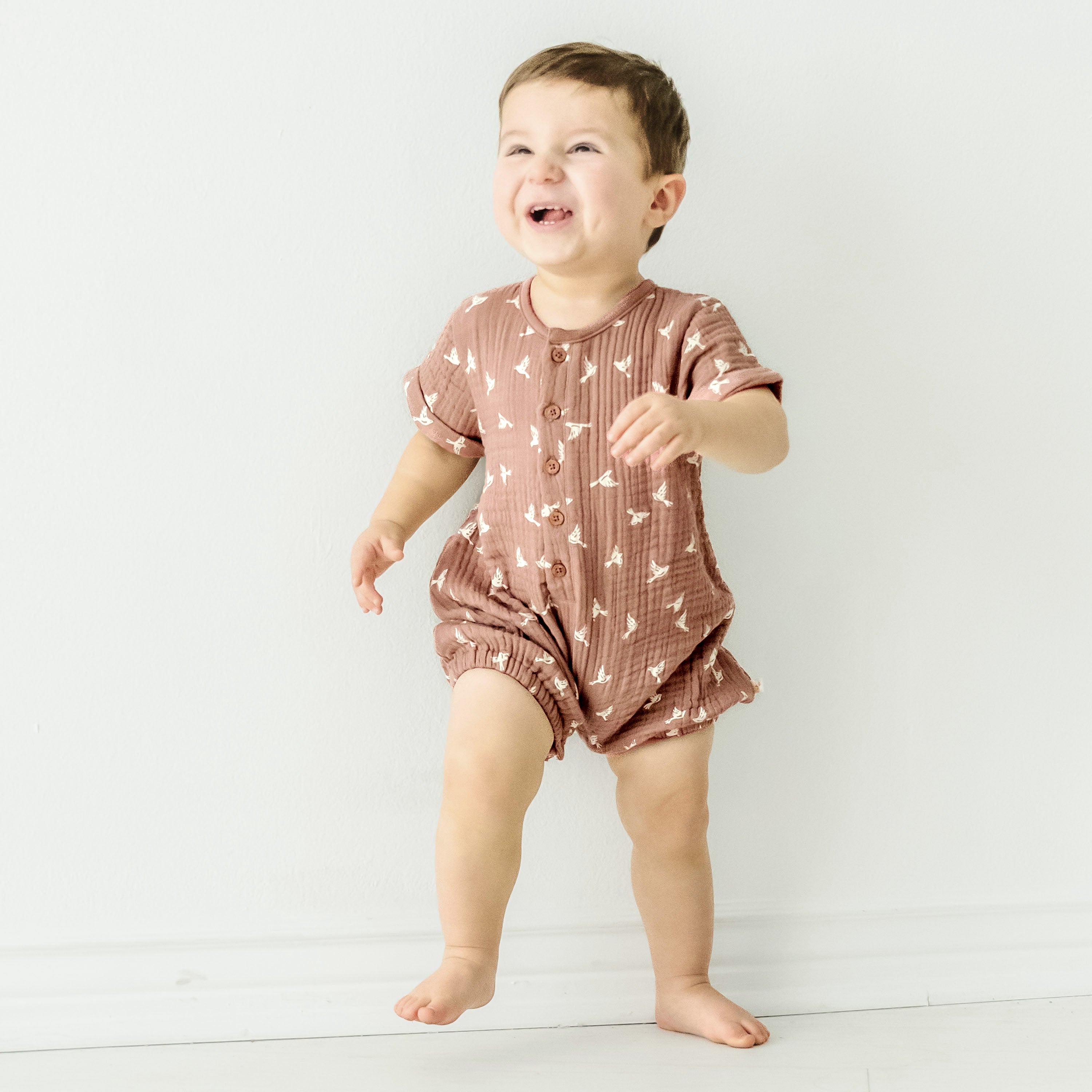 A joyful baby girl in a Makemake Organics Organic Muslin Short Bubble Romper - Flock standing barefoot against a white background, laughing and looking to the side.
