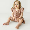 A baby girl with curly blonde hair, wearing a Makemake Organics Organic Muslin Button Flutter Dress in Flock, sits on a plain white background, looking surprised with wide eyes.