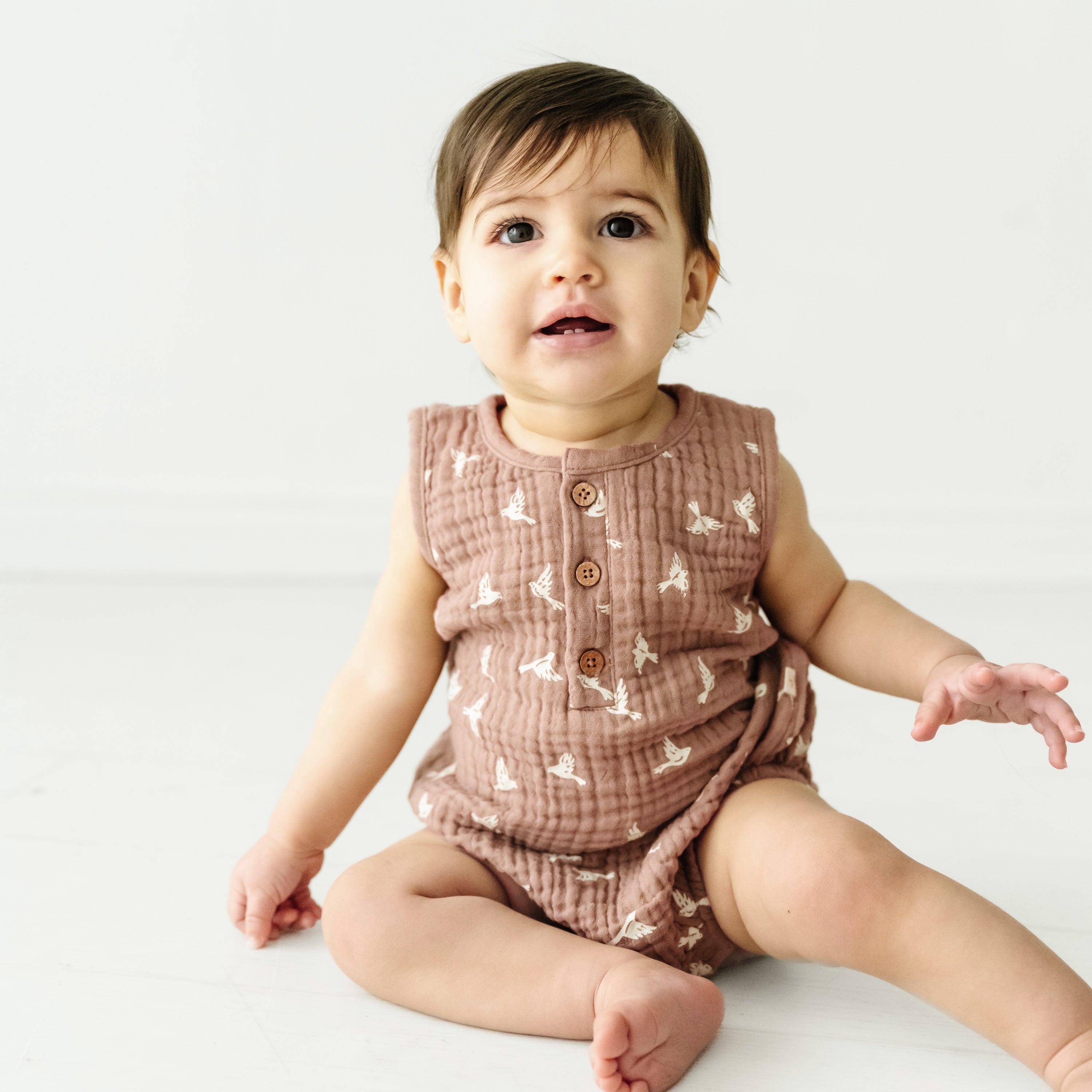 A toddler with brown hair and large eyes sits on a white surface, wearing a Makemake Organics Organic Muslin Bubble Onesie in Flock, with tiny bird patterns, looking slightly upwards with an inquisitive expression.