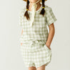 A young toddler with braided hair wearing a Makemake Organics Organic Muslin Top and Shorts 2 Piece Set - Gingham stands against a plain background.