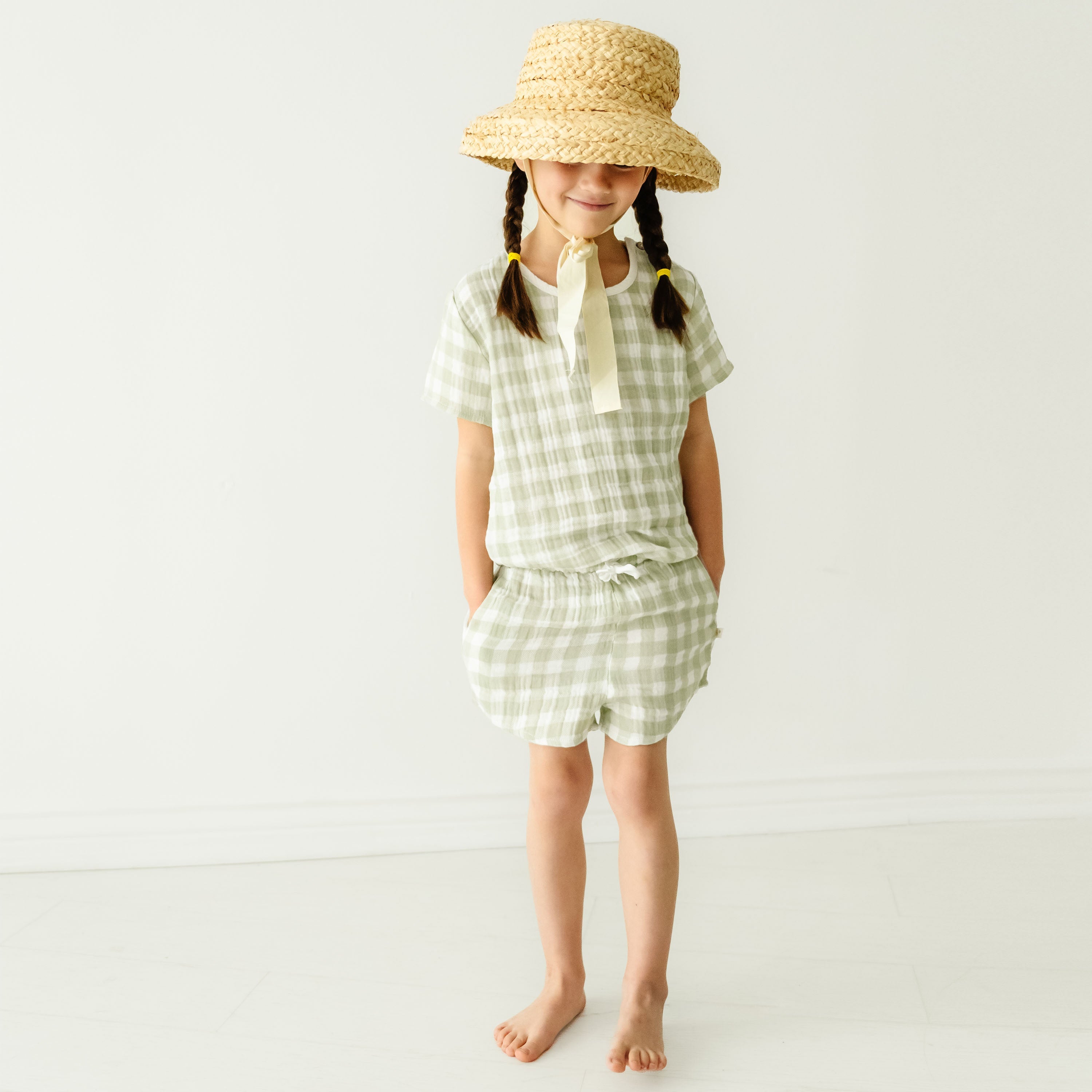 A young girl stands barefoot in a light-filled room, wearing a breezy Makemake Organics gingham dress and a wide-brimmed straw hat covering her face. Her braided hair is visible under the hat.