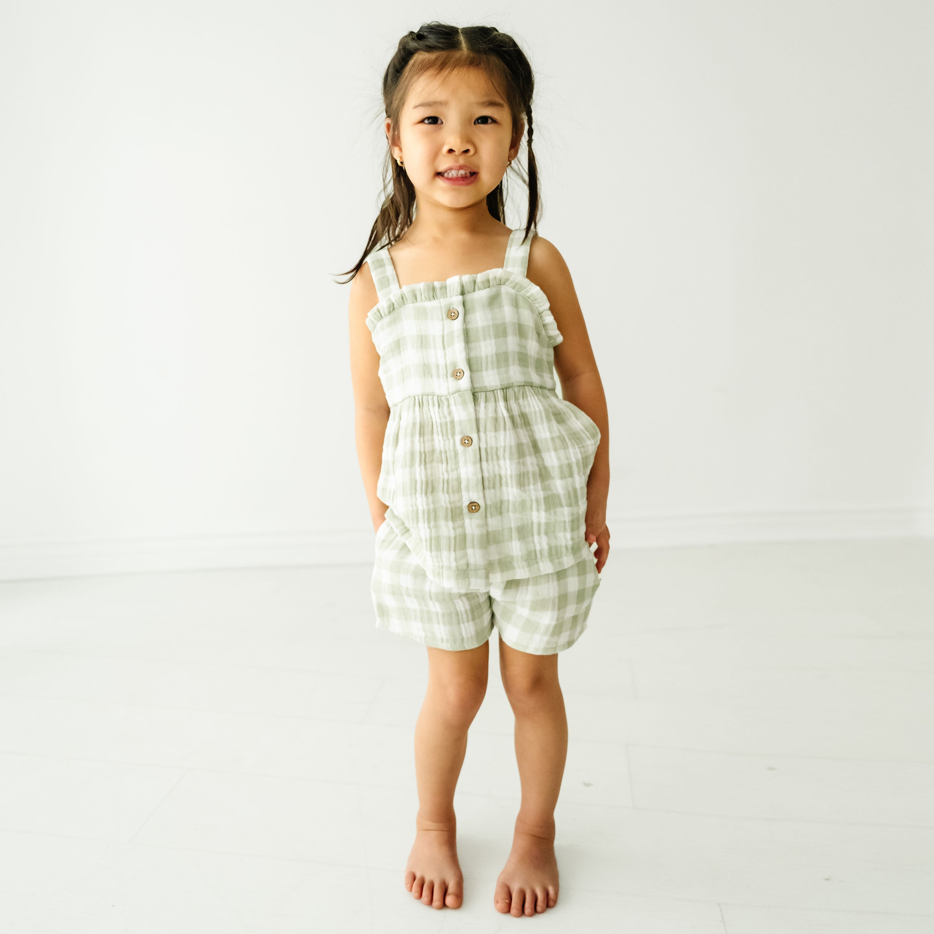 A young boy standing in a bright room, wearing a Makemake Organics Organic Muslin Peplum Top and Shorts Set - Gingham, smiling gently at the camera.