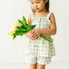 A young toddler in a Makemake Organics Organic Muslin Peplum Top and Shorts Set - Gingham holds a bouquet of white tulips, looking down at them thoughtfully against a white background.