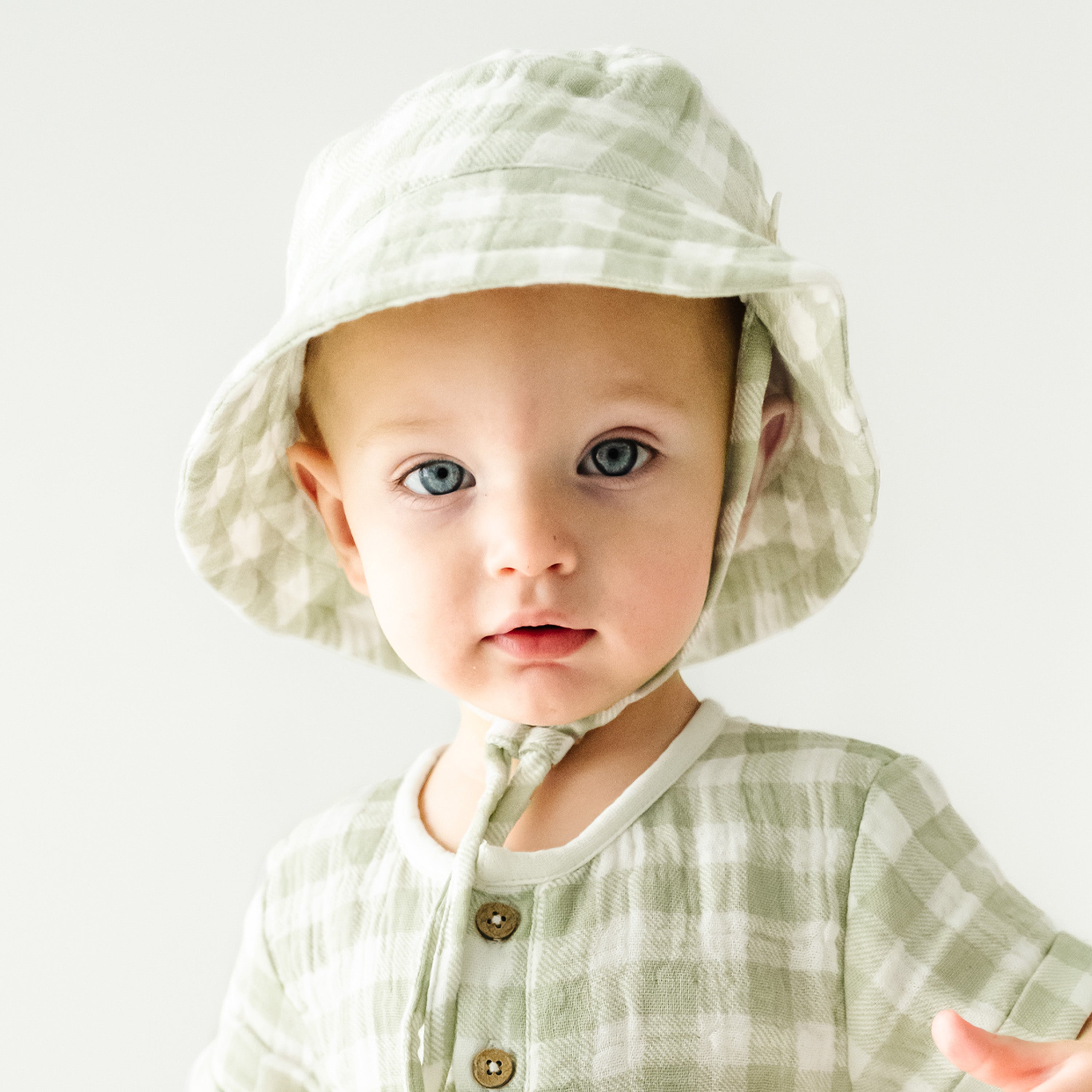 A baby with blue eyes wearing a light green checkered outfit and an Organic Muslin Bucket Sun Hat in Gingham by Makemake Organics, looking directly at the camera with a subtle expression.
