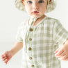 A toddler girl with blue eyes wearing a Makemake Organics Organic Muslin Short Bubble Romper - Gingham outfit and a beige sun hat, standing against a white background and looking slightly surprised.