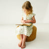 A young girl with curly hair sits on a wicker stool, reading a book attentively. She wears an Organic Muslin Button Flutter Dress - Gingham from Makemake Organics and is barefoot. The background is a plain white.