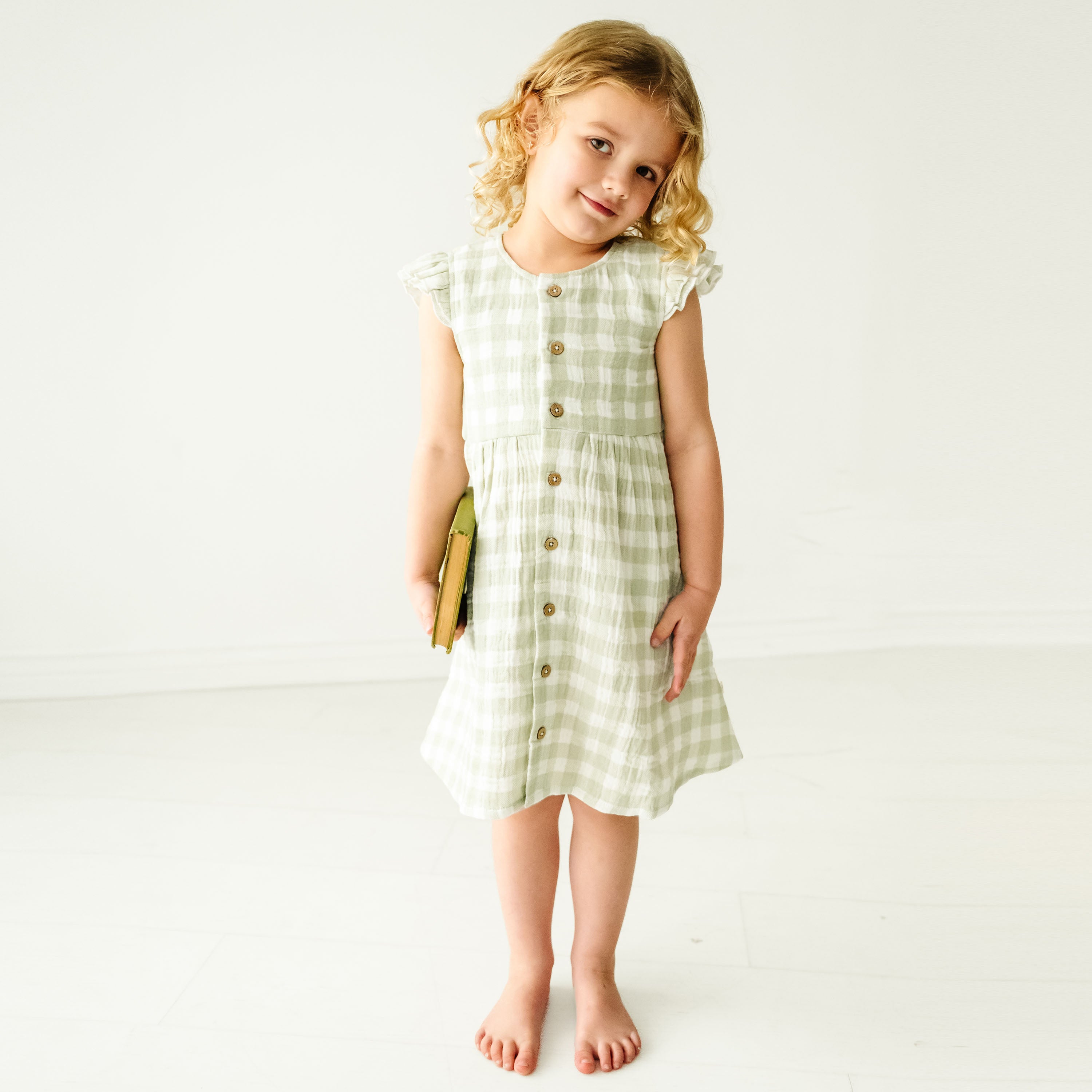 A young boy with curly hair, wearing a Makemake Organics Organic Muslin Button Flutter Dress - Gingham, standing confidently in a plain white studio, holding a book under his arm.