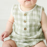 A toddler wearing a Makemake Organics Organic Muslin Bubble Onesie in Gingham sits on a neutral background, highlighting chubby arms and detailed wooden buttons on the outfit.