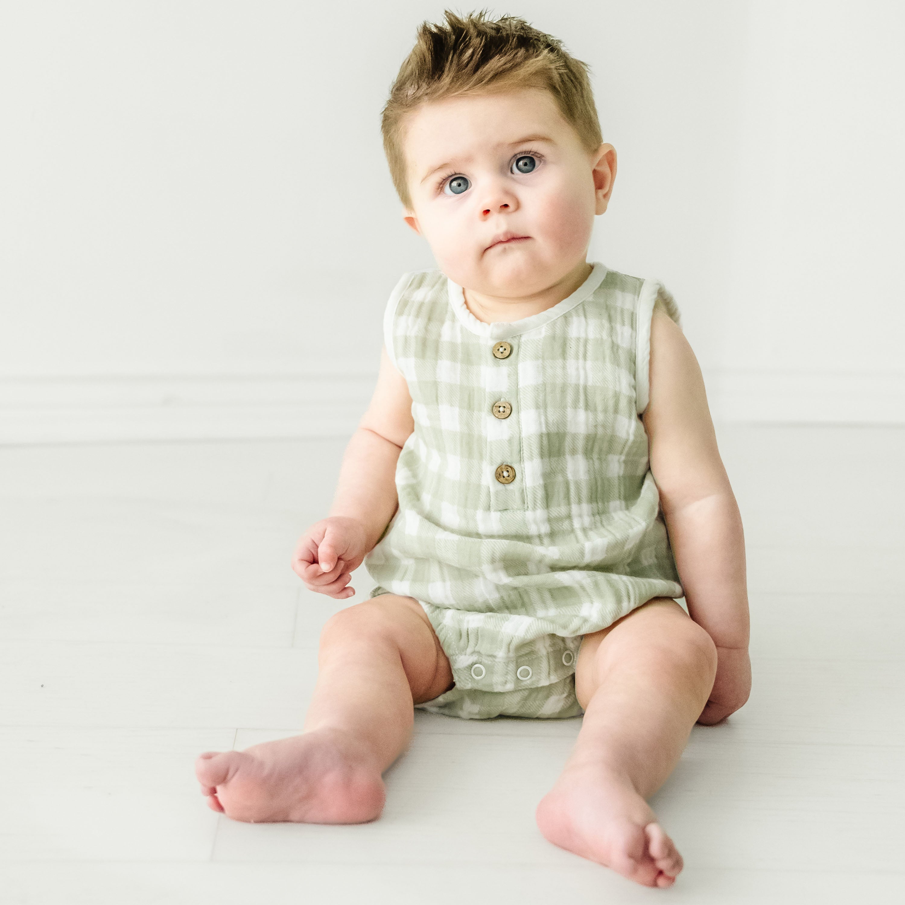 A toddler with wide eyes and tousled hair sits on a white floor, wearing a Makemake Organics Organic Muslin Bubble Onesie in Gingham with button details.