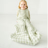 A young toddler with blonde hair sits on the floor wearing a Makemake Organics Muslin Wearable Blanket - Gingham. The child's expression is calm and gentle, against a soft white background.