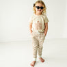 A young toddler with curly blonde hair wearing sunglasses, an Organic Crew Neck Tee - Here Comes The Sun by Makemake Organics, and striped pants, standing in a bright room, smiling.
