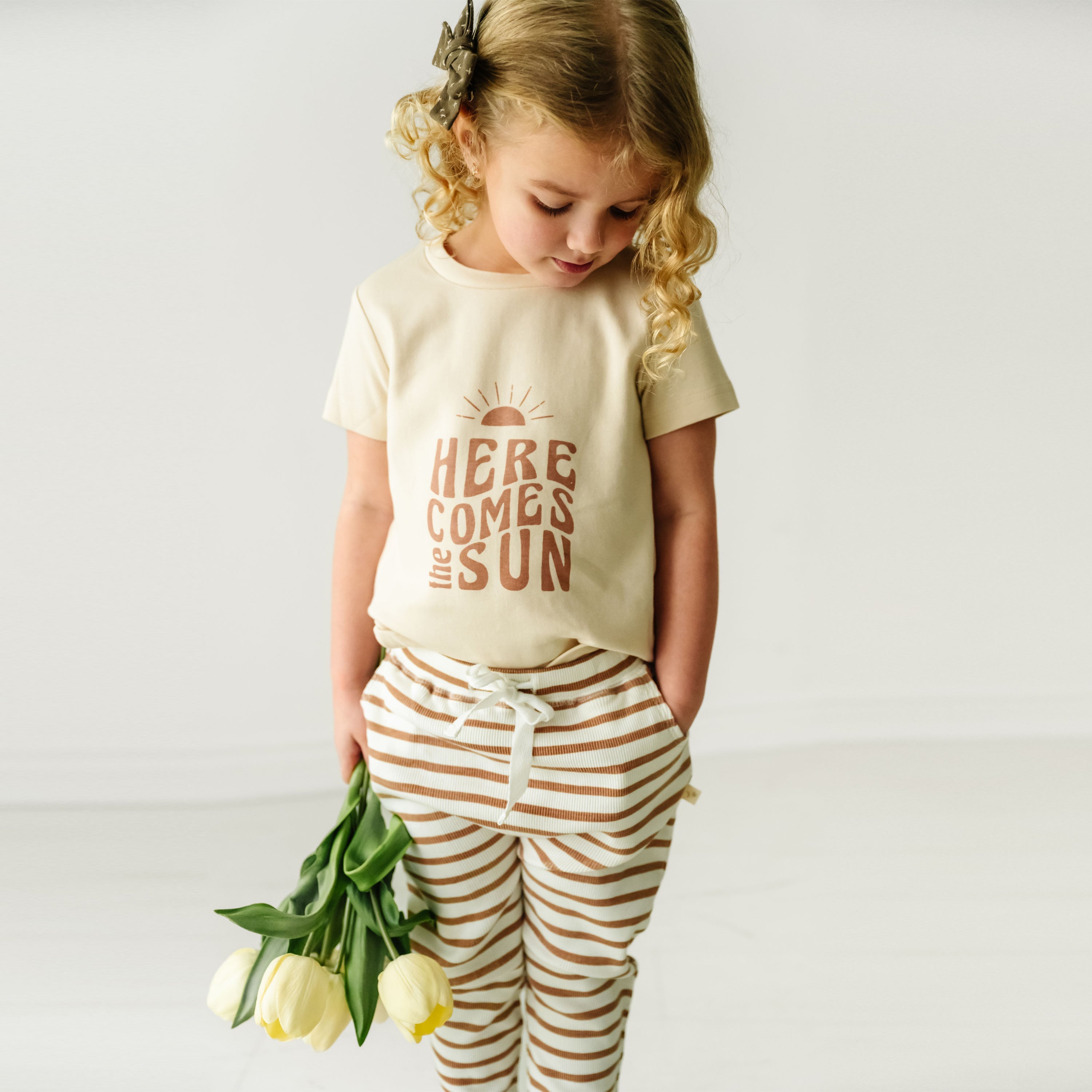 A toddler with curly blonde hair, wearing a Makemake Organics Organic Crew Neck Tee - Here Comes The Sun and striped pants, looks down at a bunch of yellow tulips she is holding.