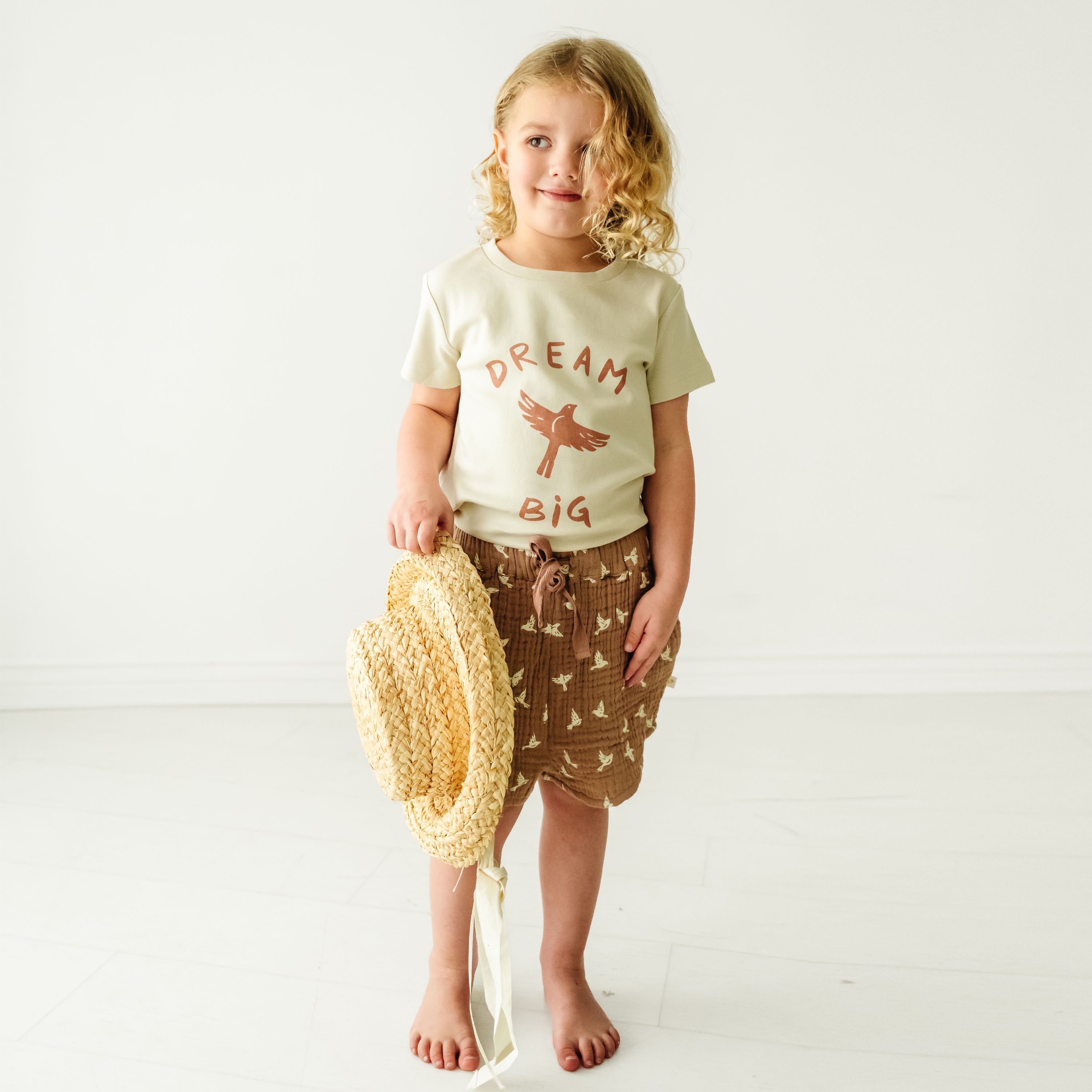 A young toddler with curly hair, wearing a cream Organic Crew Neck Tee - Dream Big t-shirt and brown shorts, stands holding a straw hat against a plain white background by Makemake Organics.