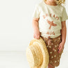 A toddler holding a straw hat stands wearing a Organic Crew Neck Tee - Dream Big by Makemake Organics, with a bird design, paired with patterned shorts. The background is bright and neutral.