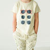 A young toddler in a Organic Crew Neck Tee - Shades by Makemake Organics with a colorful sunglasses print and dotted pants stands against a white background.