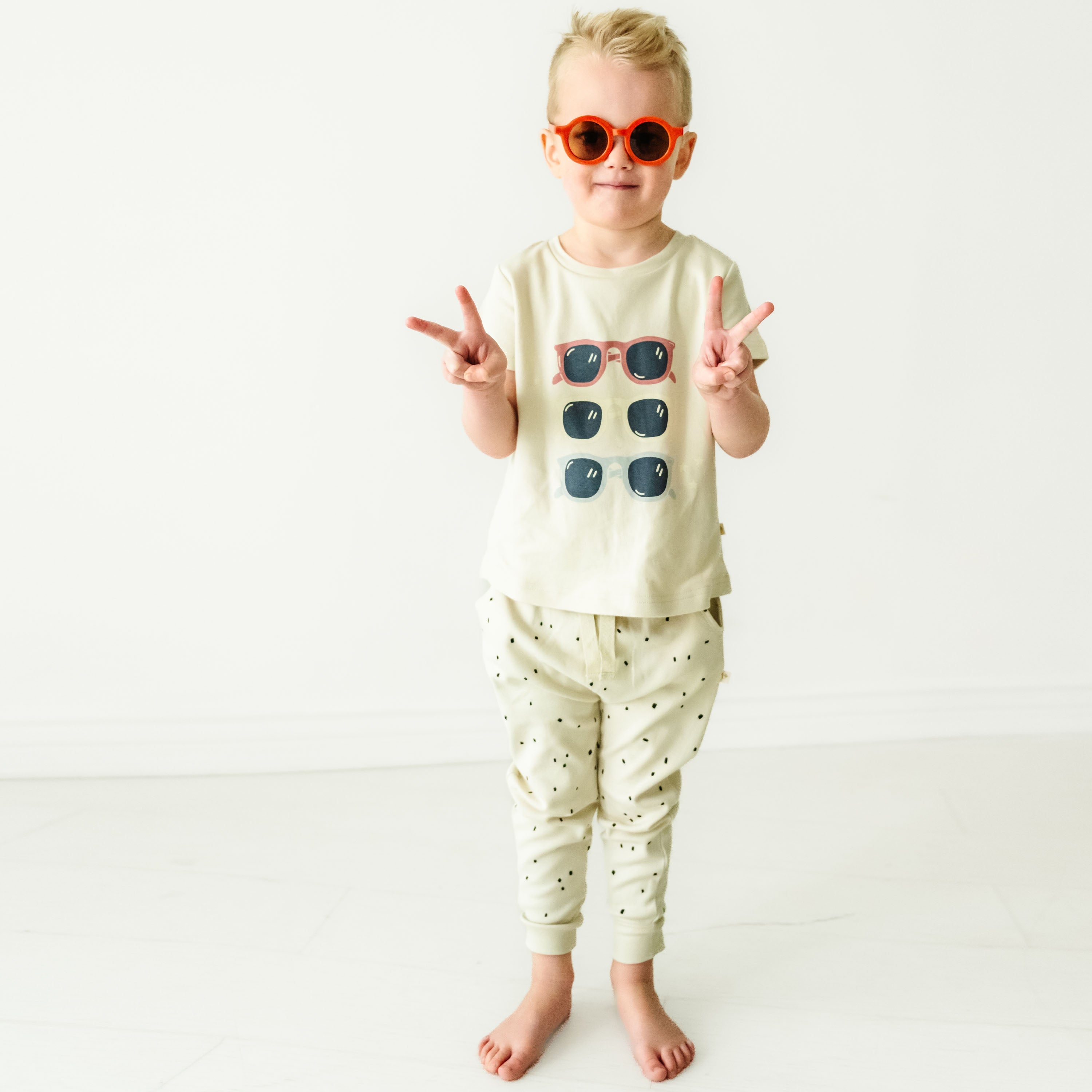 A baby wearing sunglasses and a Organic Crew Neck Tee - Shades gives a peace sign, standing barefoot on a light background.