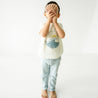 A toddler stands in a light-filled room, covering their face with a large seashell, wearing an Organic Crew Neck Tee - Ride The Waves t-shirt and pale blue pants.