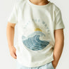 A toddler in an Organic Crew Neck Tee - Ride The Waves by Makemake Organics, paired with light blue shorts, against a neutral background.
