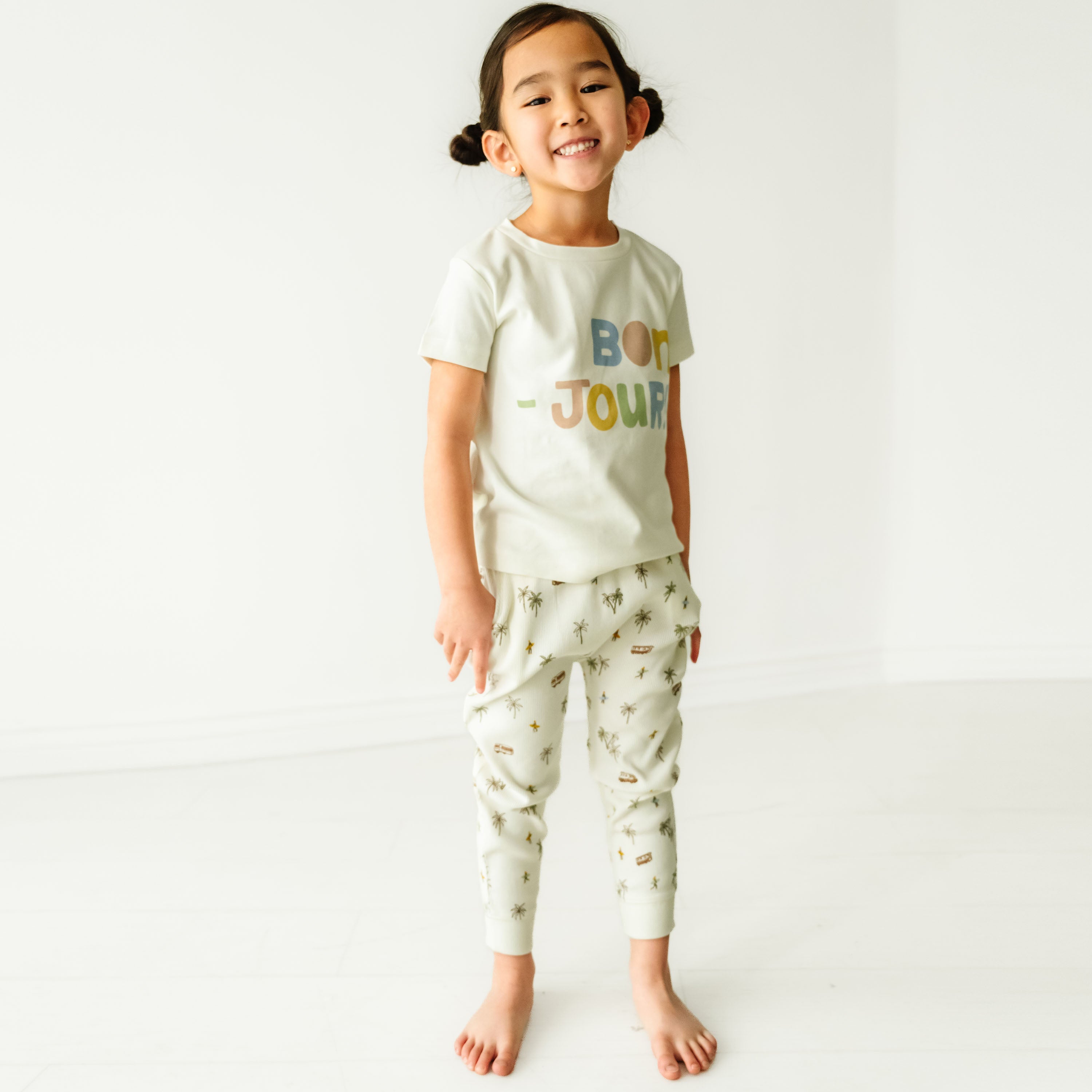 A young toddler with pigtails, smiling and wearing a white Makemake Organics Organic Crew Neck Tee - Bonjour t-shirt and patterned pants, standing in a bright, white room.