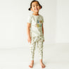 A young toddler with pigtails, smiling and wearing a white Makemake Organics Organic Crew Neck Tee - Bonjour t-shirt and patterned pants, standing in a bright, white room.