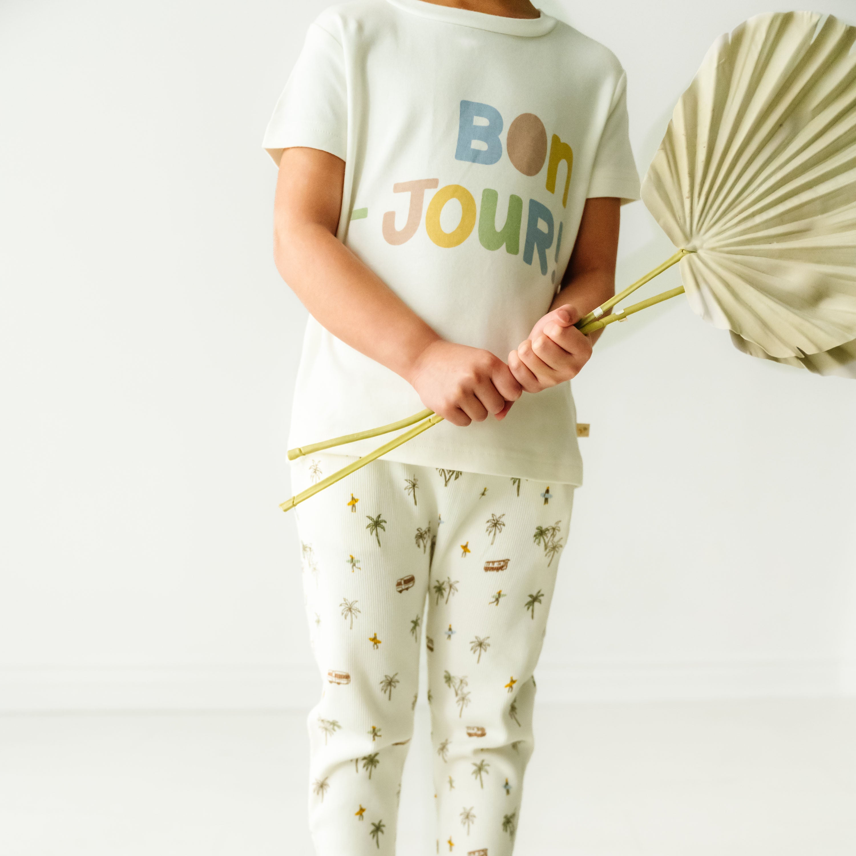 A toddler in pale pajamas printed with icons and the phrase "bon jour" on the shirt, holding a folded light-colored umbrella, standing against a white background in Makemake Organics' Organic Crew Neck Tee - Bonjour.