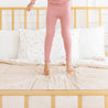 A child in pink pajamas is playfully jumping on a bed with floral bedding and a wooden frame, with only the lower half of their body visible, using the Makemake Organics Organic Cotton Sheet Set in Polka Dots.