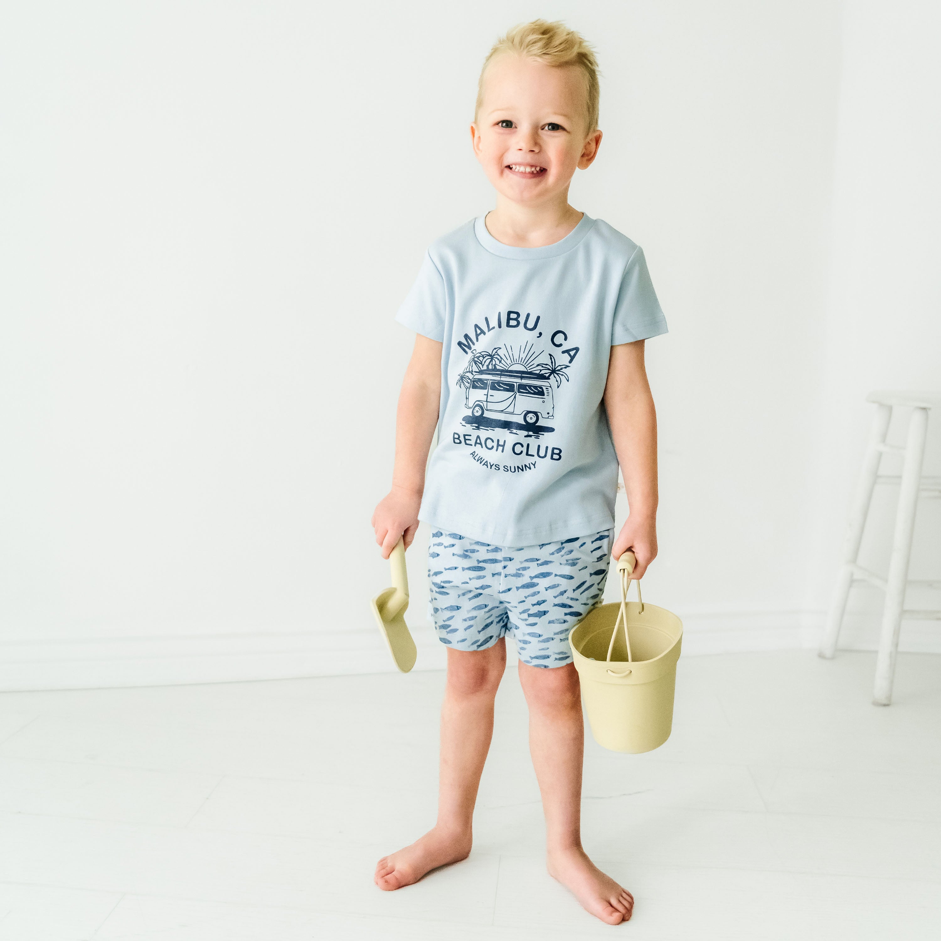 A joyful toddler boy standing in a white studio, wearing a Malibu Beach Club Organic Crew Neck Tee and blue patterned shorts, holding a yellow sand scoop and a pale yellow bucket.