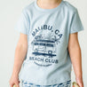 A toddler wearing a light blue Organic Crew Neck Tee with a graphic of a van, palm trees, and the words "malibu, ca beach club beach days sunny" printed on it. The background is plain.