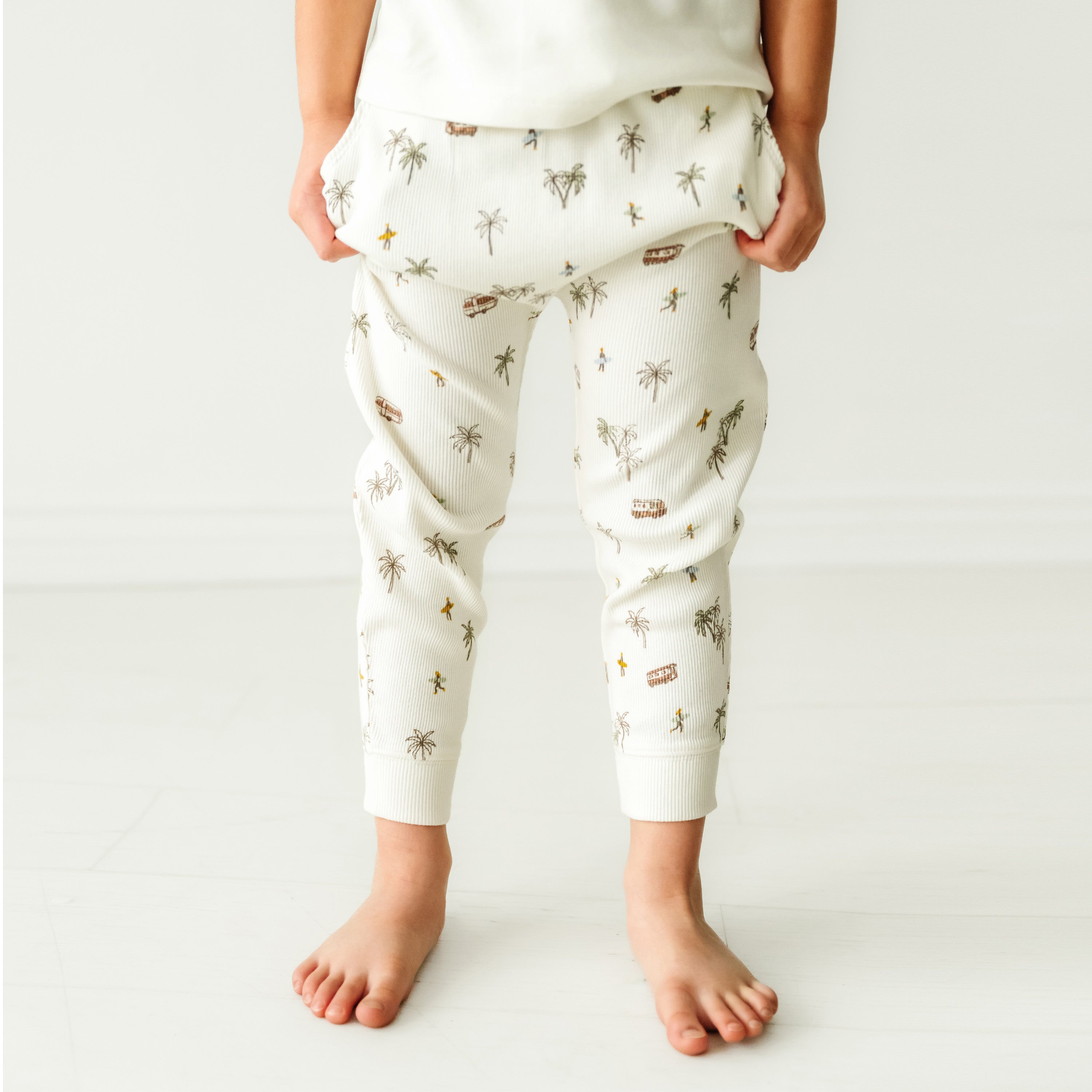 A toddler standing barefoot wearing white pajamas with a whimsical pattern of stars and subtle earth-toned designs. The focus is on the Organic Harem Pants - Malibu by Makemake Organics and the child's lower torso and legs.