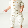 A baby boy in a white Makemake Organics Organic Short Sleeve Button Romper adorned with palm tree designs takes cautious steps in a bright room. The outfit also features buttons and the child is barefoot.