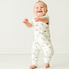 A joyful baby girl smiling and taking a step forward, dressed in a white Organic Short Sleeve Button Romper with a beach-themed pattern, in a bright, neutral-toned room from Makemake Organics.