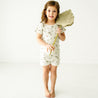 A young child stands barefoot in a white room, smiling and holding a large, leaf-shaped fan. she wears a light-colored dress adorned with small botanical prints. She is wearing the Organic Tee and Shorties Set from Makemake Organics - Malibu.