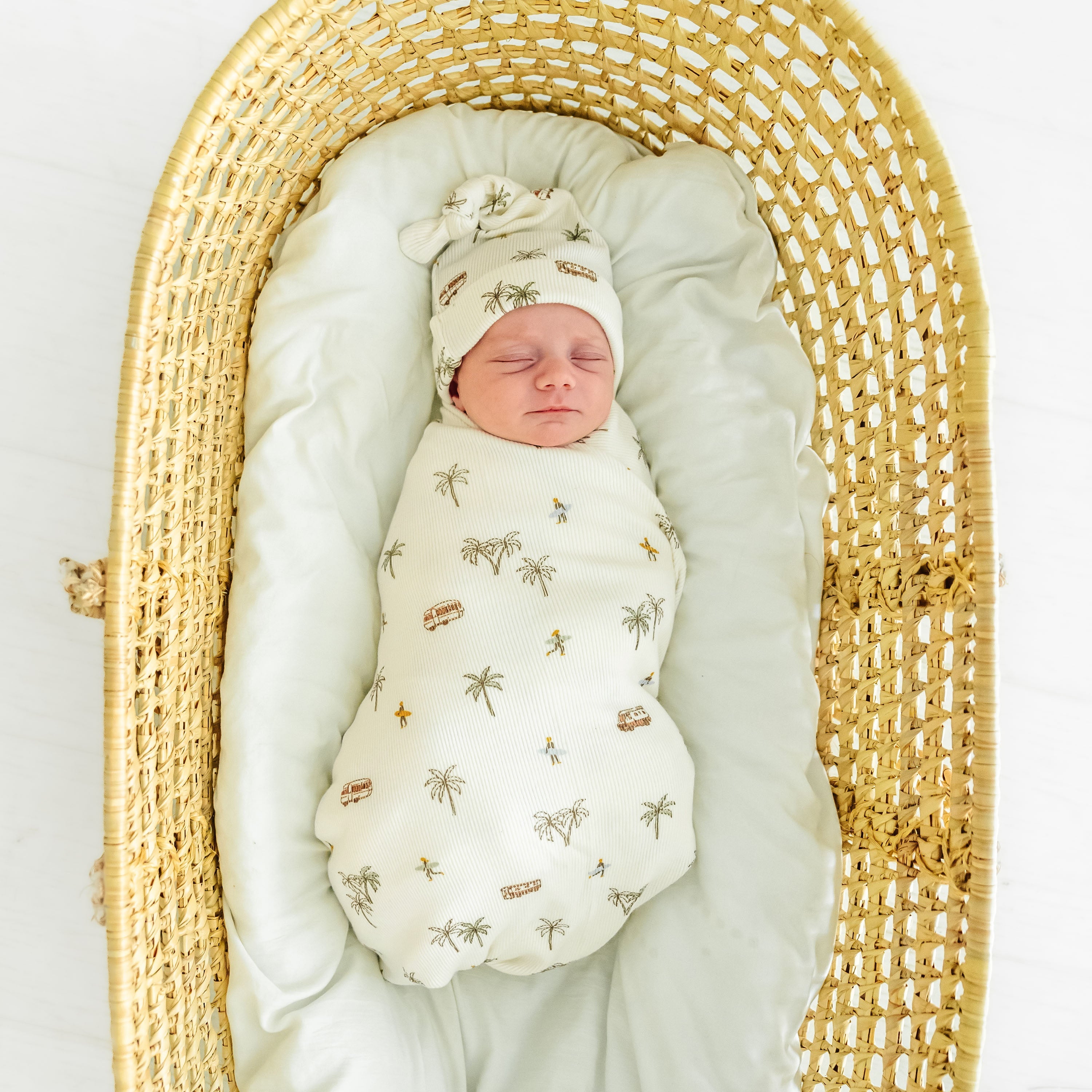 A newborn baby swaddled in an Organic Swaddle Blanket & Hat from Makemake Organics, sleeping in a padded, oval-shaped wicker bassinet on a white background.