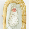 A newborn baby swaddled in an Organic Swaddle Blanket & Hat from Makemake Organics, sleeping in a padded, oval-shaped wicker bassinet on a white background.